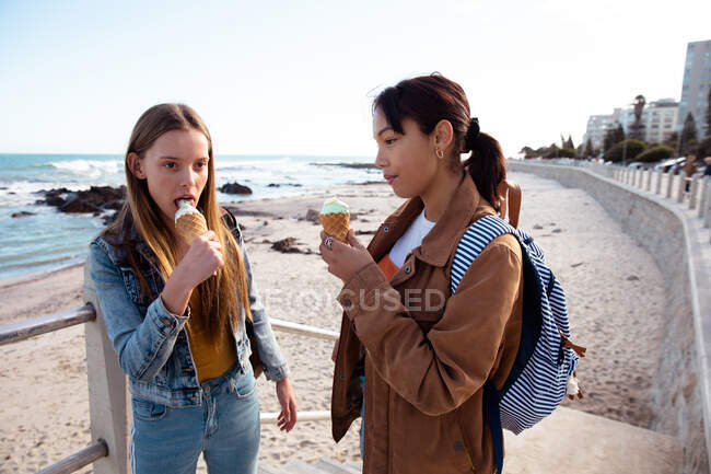 Vista frontal de un caucásico y una mezcla de chicas de raza disfrutando del tiempo juntos en un día soleado, comiendo helado, de pie en un paseo marítimo. - foto de stock