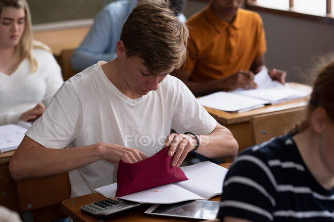 Вид сбоку на мальчика-белого подростка в школьном классе, сидящего за столом, ищущего ручку в своем пенал-кейсе, с одноклассниками-подростками, сидящими за партами, работающими на заднем плане — стоковое фото