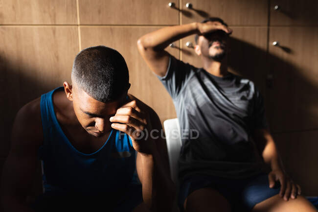 Zwei gemischte männliche Fußballspieler in Sportklamotten sitzen während einer Spielpause in der Umkleidekabine und ruhen sich enttäuscht aus. — Stockfoto