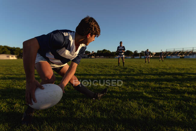 Vista laterale di un adolescente caucasico giocatore di rugby maschile che indossa una striscia di squadra blu e bianca, pronto a passare la palla da rugby ai compagni di squadra in piedi sullo sfondo, su un campo di gioco. — Foto stock
