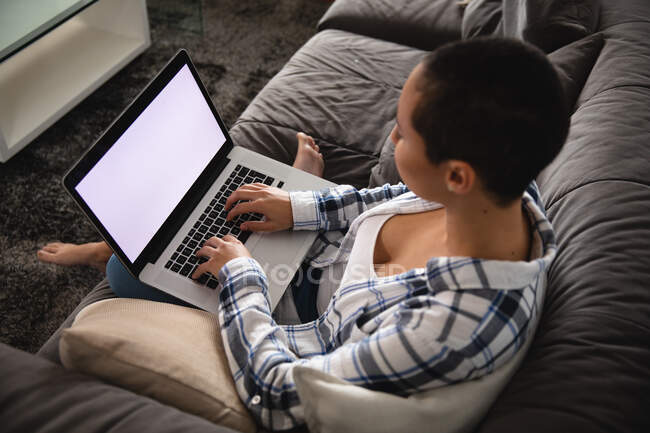 Vista de ángulo alto de la mujer de raza mixta que se relaja en casa, sentado en un sofá con los pies desnudos, utilizando una computadora portátil - foto de stock