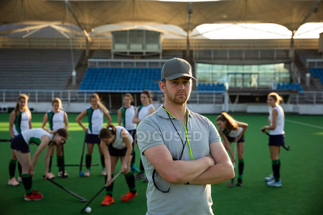 Портрет кавказского тренера по хоккею на траве, подготовка команды к матчу, стоя на хоккейном поле, глядя в камеру, со своей командой, играющей с мячом на заднем плане в солнечный день — стоковое фото