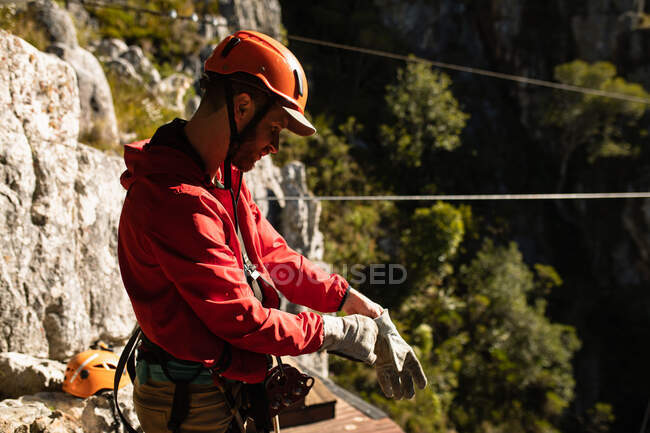Seitenansicht des kaukasischen Mannes, der die Zeit in der Natur genießt, Ausrüstung mit Reißverschluss trägt, Handschuhe anzieht, sich auf Reißverschluss vorbereitet, an einem sonnigen Tag in den Bergen — Stockfoto