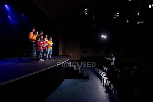 Vista lateral de um grupo multi-étnico de coristas adolescentes do sexo masculino e feminino segurando partituras e cantando em pé no palco de um teatro escolar durante os ensaios para uma performance — Fotografia de Stock