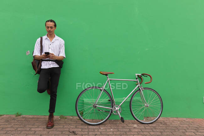Vista frontal de un hombre de raza mixta con largas rastas fuera y alrededor de la ciudad en un día soleado, de pie en la calle, apoyado contra la pared verde, utilizando un teléfono inteligente, con su bicicleta de pie junto a él. - foto de stock
