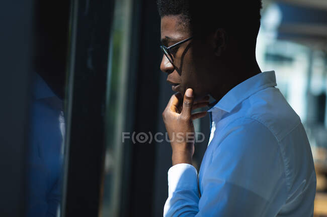 Un hombre de negocios afroamericano con el pelo corto, con una camisa blanca y gafas de pie dentro de un café y mirando a la ventana tocándose la barbilla - foto de stock