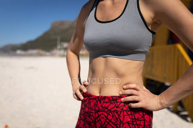 Вид спереду середня секція жінки, в спортивному одязі, стоїть руками на стегнах на пляжі, розслабляється після бігу . . — стокове фото