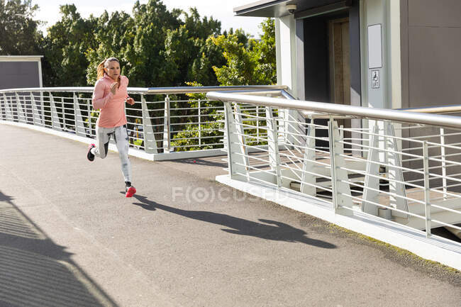 Боковой вид спортивной белой женщины с длинными темными волосами, тренирующейся в городском парке в солнечный день с голубым небом, бегущей. — стоковое фото