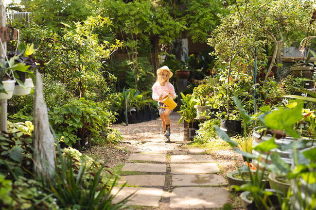Una ragazza caucasica con lunghi capelli biondi che si gode il tempo in un giardino soleggiato, esplorando, correndo, tenendo in mano un annaffiatoio, indossando un cappello di paglia — Foto stock