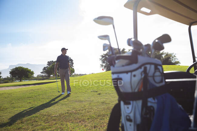 Vorderansicht eines kaukasischen Mannes auf einem Golfplatz an einem sonnigen Tag mit blauem Himmel, der neben einem Golfwagen einen Golfschläger hält — Stockfoto