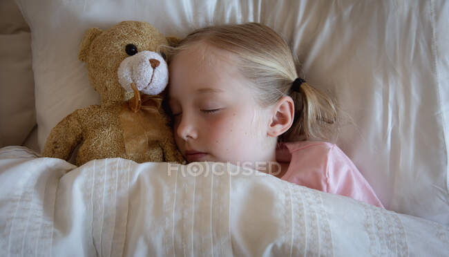 Вид спереди под высоким углом на белую девушку, наслаждающуюся свободным временем дома, спящую в своей постели, рядом со своим плюшевым мишкой — стоковое фото
