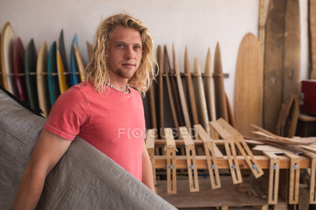 Creatore caucasico di tavole da surf da uomo che lavora nel suo studio, con una tavola da surf ricoperta da una custodia grigia, con tavole da surf in una cremagliera sullo sfondo. — Foto stock
