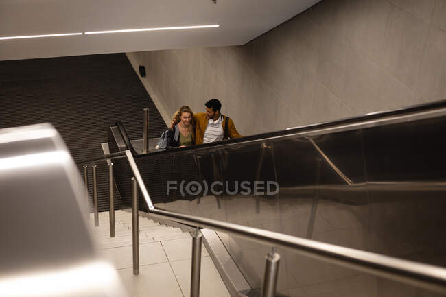 Vista frontal de ángulo alto de una pareja caucásica en la ciudad, subiendo en la estación de metro con una escalera mecánica, sonriendo y abrazando. - foto de stock