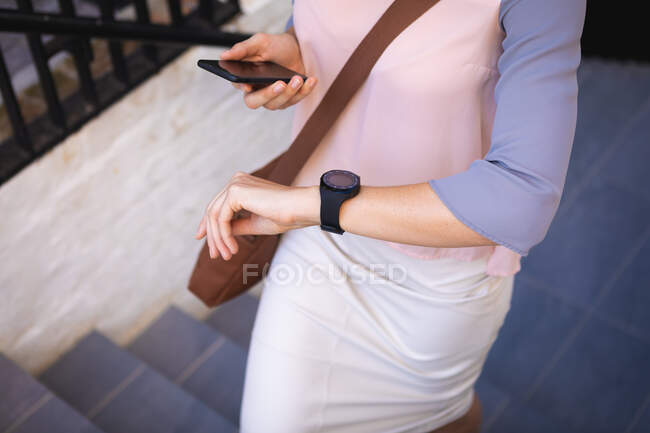 Mujer de negocios en movimiento en un día soleado, caminando por las escaleras, sosteniendo su teléfono inteligente y comprobando un reloj inteligente - foto de stock