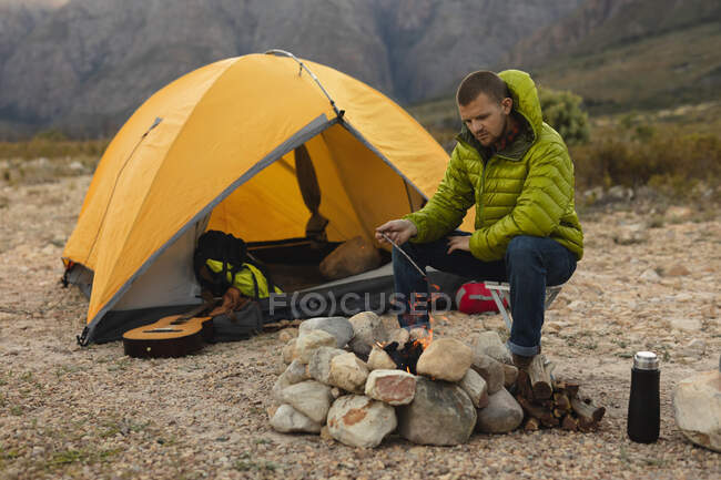 Vista lateral de um homem caucasiano se divertindo em uma viagem às montanhas, sentado perto de uma fogueira, observando o fogo — Fotografia de Stock