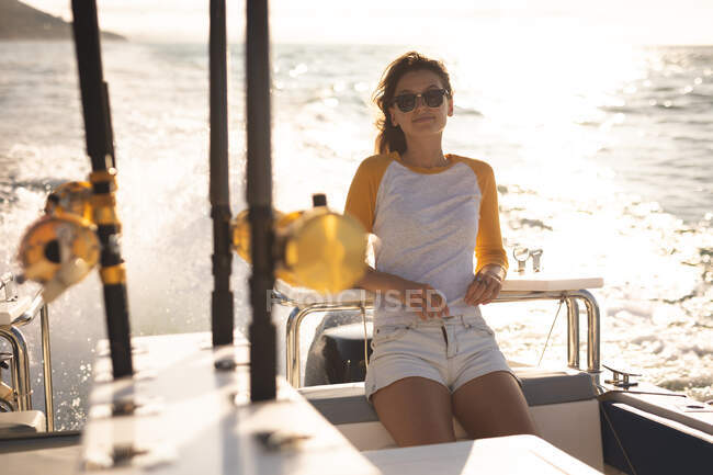 Retrato de una adolescente caucásica disfrutando de su tiempo de vacaciones al sol junto a la costa, parada en un barco, inclinada, relajada, mirando a la cámara y sonriendo - foto de stock