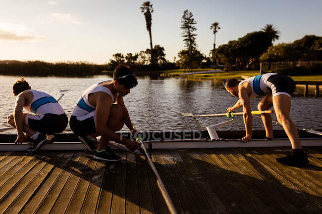 Side view of three Caucasian male rowers preparing a rowing boat before rowing, standing on a jetty on the river, holding oars — Stock Photo