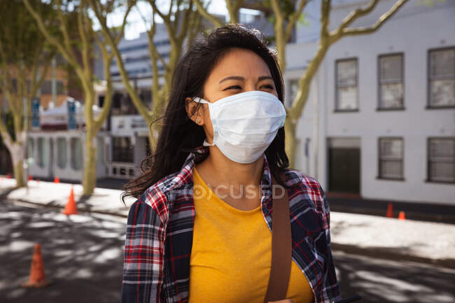 Side view of a mixed race woman with long dark hair out and about in the city streets during the day, wearing a face mask against air pollution and coronavirus, standing in a city street with buildings in the background. — Stock Photo