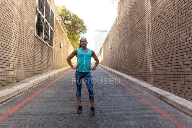 Переду видніється спортивна кавказька жінка з довгим темним волоссям, що вправляється в міській місцевості, стоячи з руками на стегнах.. — стокове фото