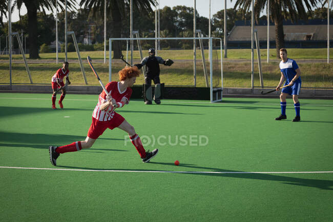 Side view of a teenage Caucasian male field hockey player in action on the pitch, preparing to hit a ball during a hockey game on a sunny day — Stock Photo
