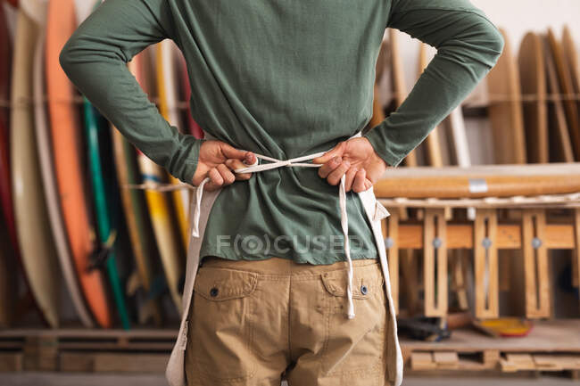 Vista posteriore del costruttore di tavole da surf maschile nel suo studio, indossando un grembiule protettivo, allacciando i lacci, con tavole da surf in un rack sullo sfondo. — Foto stock
