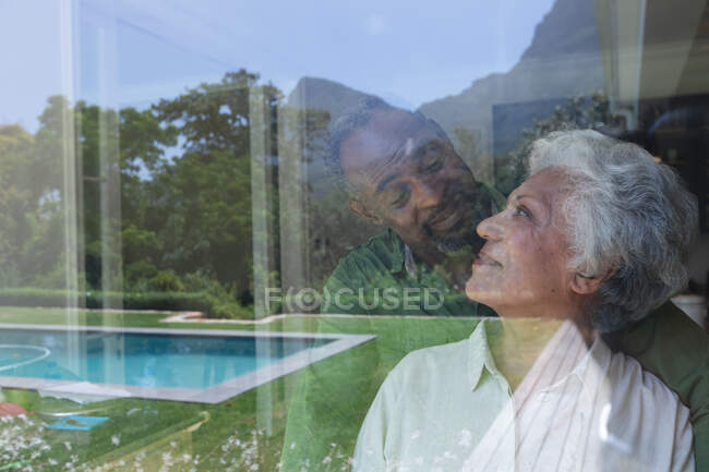 Feliz pareja afroamericana jubilada jubilada en casa, mirándose y sonriendo, reflejada en una ventana con vistas a su jardín con piscina, pareja en casa aislándose juntos durante la pandemia de coronavirus covid19 - foto de stock