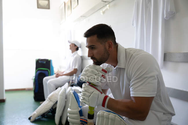 Side view close up of a mixed race male cricket player wearing whites, sitting on a bench in a changing room, propping up his head on a cricket bat, with another player sitting in behind. — Stock Photo