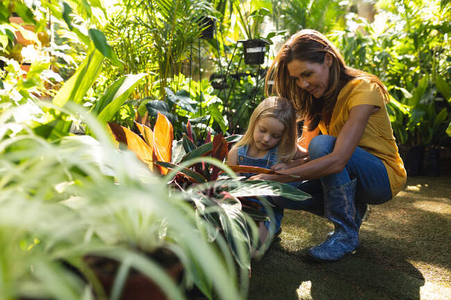 Une femme caucasienne et sa fille s'amusent ensemble dans un jardin ensoleillé, regardant les plantes ensemble et touchant leurs feuilles et leurs fleurs — Photo de stock