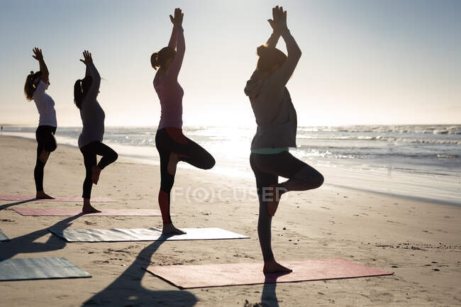 Rear view of a multi-ethnic group of female friends enjoying exercising on a beach on a sunny day, practicing yoga, standing in tree position. — Stock Photo