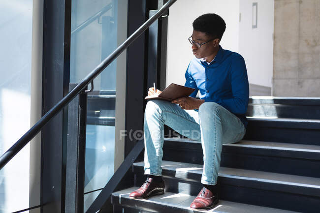 Un uomo d'affari afroamericano con i capelli corti scuri, indossa una camicia blu e occhiali, lavora in un ufficio moderno, si siede sulle scale e scrive appunti — Foto stock