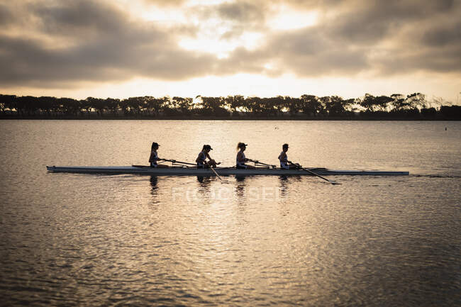 Vista lateral de un equipo de remo de cuatro mujeres caucásicas entrenando en el río, remando en una concha de carreras al amanecer, con la luz del sol reflejada en las ondas del agua en primer plano - foto de stock