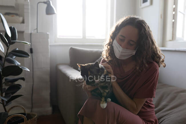 Una donna caucasica passa del tempo a casa, giocando con il suo gatto, indossando una maschera. Stile di vita a casa isolante, distanza sociale in isolamento di quarantena durante il coronavirus covid 19 pandemia. — Foto stock