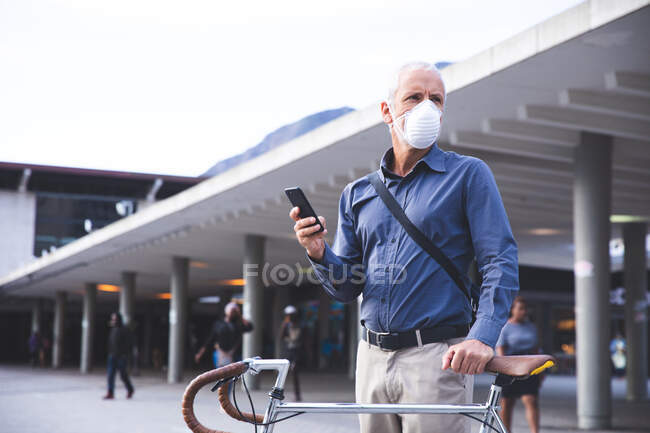 Hombre caucásico mayor por las calles de la ciudad durante el día, usando una máscara facial contra el coronavirus, covid 19, moviendo su bicicleta y usando un teléfono inteligente. - foto de stock