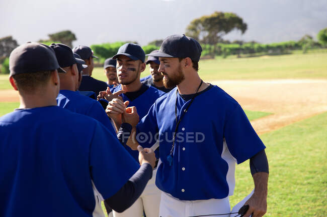 Вид спереди на многонациональную группу бейсболистов-мужчин, стоящих на поле после тренировки и встречающихся со своим тренером и товарищами по команде в солнечный день — стоковое фото