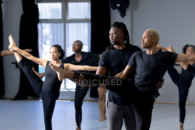 Frontansicht eines modernen Tänzers mit gemischter Rasse, der ein schwarzes Outfit trägt und einen männlichen Tänzer unterstützt, während er sich während eines Tanzkurses in einem hellen Studio hochstreckt, während andere Tänzer im Hintergrund trainieren. — Stockfoto