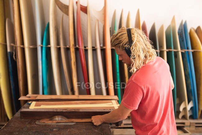 Fabricante de tablas de surf masculino caucásico trabajando en su estudio, usando auriculares protectores, cortando rayas de madera y preparándose para hacer una tabla de surf, con tablas de surf en un estante en el fondo. - foto de stock