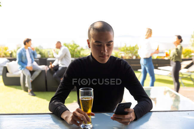 Вид спереди на смешанного гонщика, болтающегося на террасе на крыше в солнечный день, использующего смартфон и держащего стакан пива, с людьми, разговаривающими на заднем плане — стоковое фото