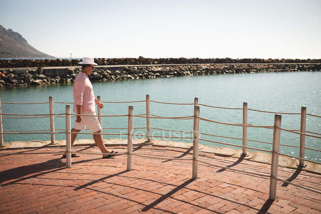 Белый мужчина, одетый в розовую рубашку и шляпу, наслаждающийся прогулкой на набережной, в солнечный день, гуляющий — стоковое фото