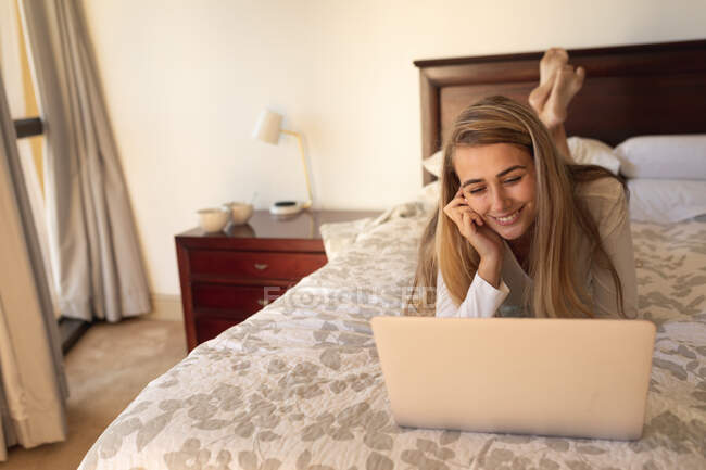Белая женщина лежит на кровати, используя ноутбук. Социальное дистанцирование и самоизоляция в карантинной изоляции. — стоковое фото