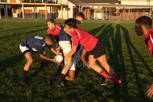 Vista lateral de dos equipos masculinos multiétnicos adolescentes de jugadores de rugby que usan sus tiras de equipo, en acción durante un partido de rugby en un campo de juego, un jugador en tira azul y blanca en posesión de la pelota - foto de stock