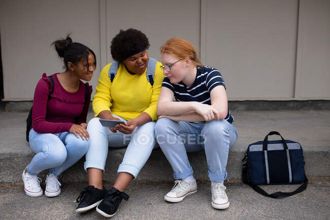 Vista frontal de um grupo multiétnico de alunas adolescentes do ensino médio com bolsas de estudo penduradas, conversando e olhando para um smartphone juntas, sentadas em degraus em seus terrenos escolares — Fotografia de Stock