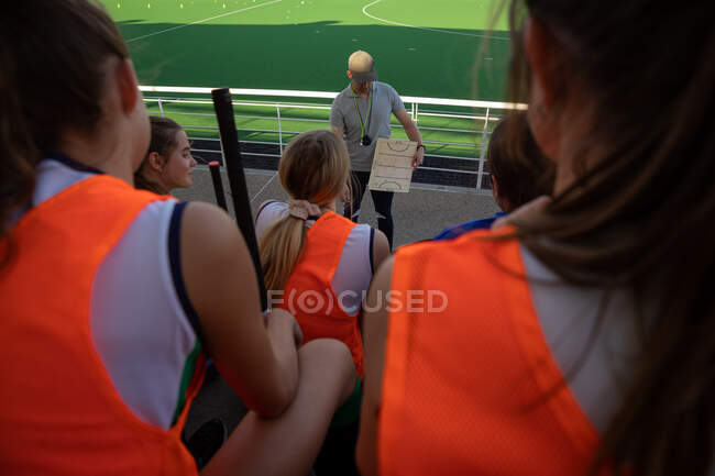 High angle rear view of a group of female Caucasian field hockey players preparing before a game, sitting on a stand, holding hockey sticks, with their Caucasian male field hockey coach standing in front of them — Stock Photo