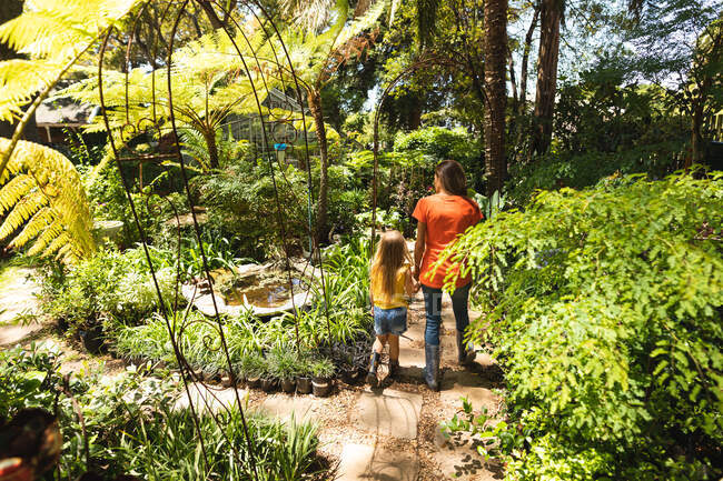 Кавказская женщина и ее дочь наслаждаются временем вместе в солнечном саду, держась за руки и идя по тропинке между растениями — стоковое фото