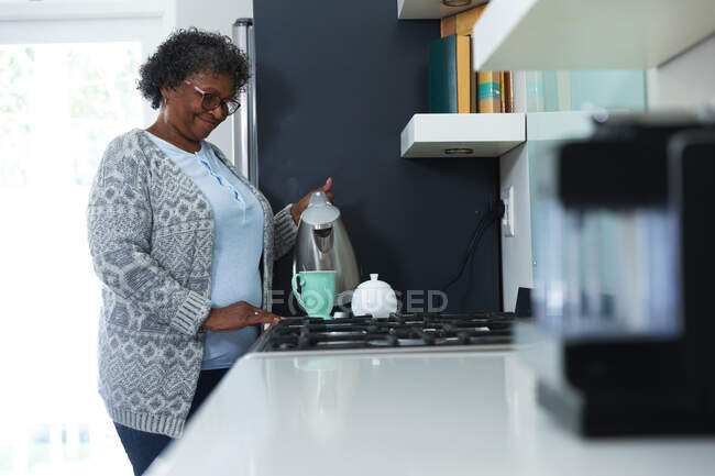 Senior mujer de raza mixta disfrutando de su tiempo en casa, distanciamiento social y auto aislamiento en cuarentena, de pie en su cocina, preparando té - foto de stock