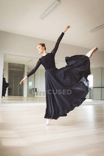 Bailarina de ballet femenina atractiva caucásica con ballet de baile de pelo rojo, vestida con un vestido largo y negro, preparándose para una clase de ballet en un estudio brillante, centrándose en su ejercicio. - foto de stock