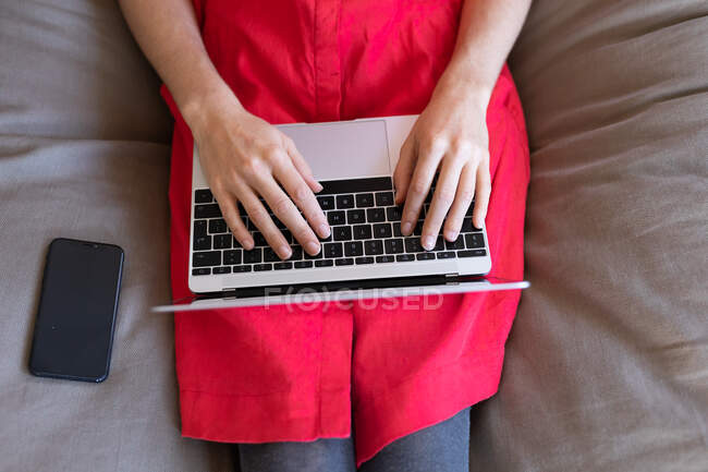 Partie médiane de la femme passant du temps à la maison, portant une robe rose, assis sur un canapé et utilisant son ordinateur portable. Distance sociale et isolement personnel en quarantaine. — Photo de stock