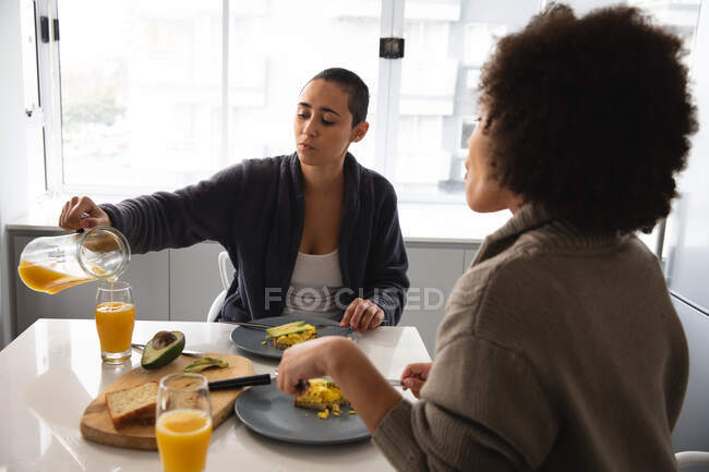 Вид спереди на женскую пару смешанной расы, отдыхающую дома, сидящую за столом на кухне, завтракающую и разговаривающую, одну наливающую стакан апельсинового сока из кувшина — стоковое фото