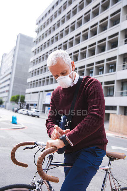 Homme caucasien âgé dehors et autour dans les rues de la ville pendant la journée portant un masque facial contre le coronavirus, covid 19, assis sur un vélo et en utilisant une montre intelligente. — Photo de stock