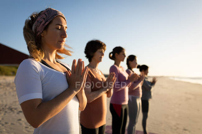 Vista laterale di un gruppo multietnico di amiche che si esercitano su una spiaggia in una giornata di sole, praticano yoga in piedi, con le mani tenute in posizione di preghiera e gli occhi chiusi. — Foto stock