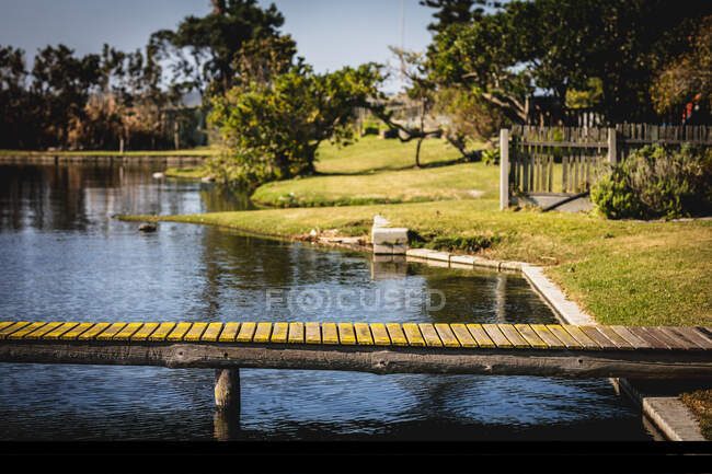 Un molo in un lago su un parco e alberi su una riva erbosa con recinzioni in una giornata di sole — Foto stock